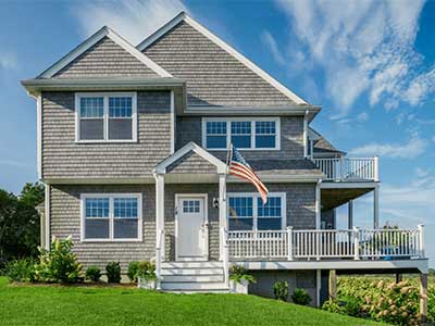 Front view of a two-story with an American flag in the front, a front porch and balcony on the side, and gray Tando siding